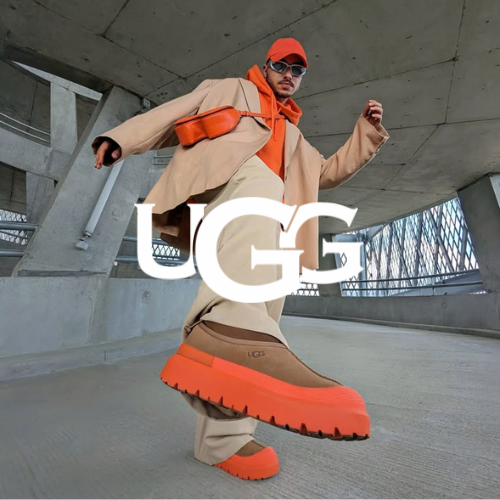 UGG Kollektion – Mann in Betonkomplex steht in braunem Outfit mit orangenen Details und hält UGG Boots in die Kamera, urbaner Lifestyle bei Closory