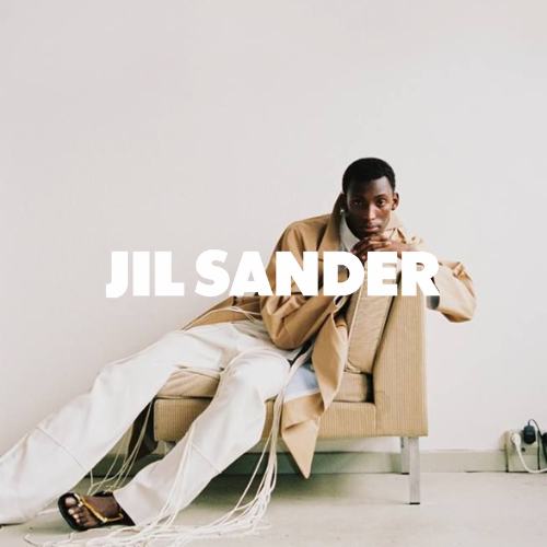 Jil Sander Kollektion – Model in weißer Hose und braunem Mantel sitzend auf gleichfarbigem Stuhl, eleganter Lifestyle bei Closory