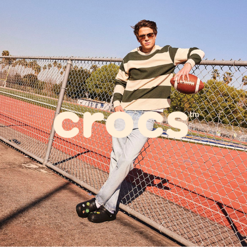 Crocs Kollektion – Mann lehnt an Zaun vor Sprintstrecke mit Crocs an, lässiger Lifestyle bei Closory